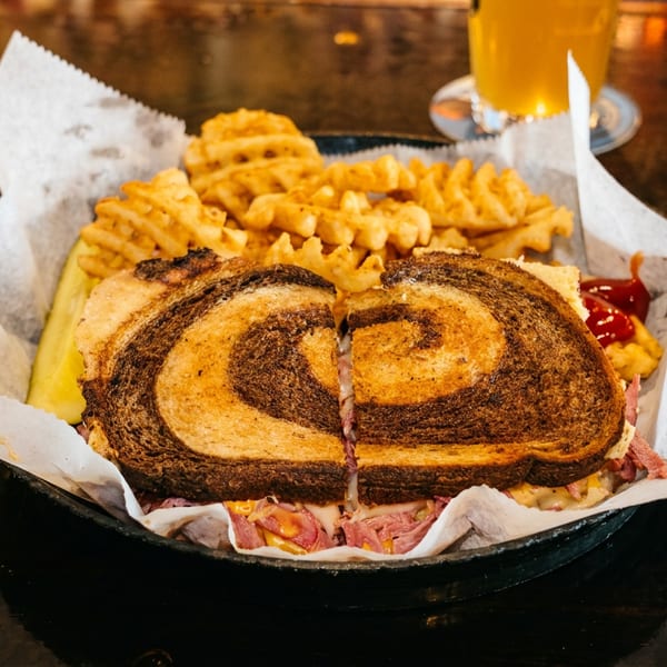 A deli sandwich on marbled rye bread with waffle fries and a pickle spear in a basket.