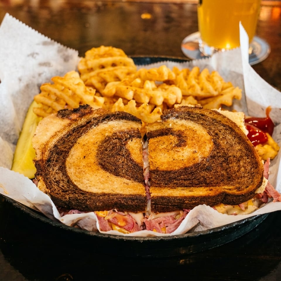 A deli sandwich on marbled rye bread with waffle fries and a pickle spear in a basket.