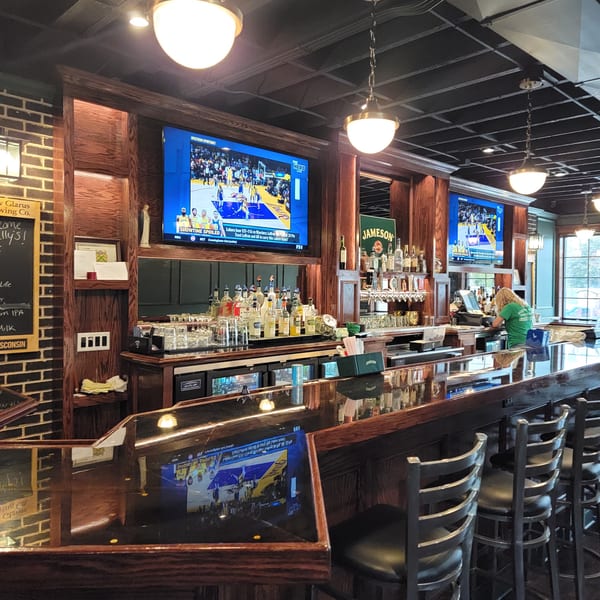 A long, dark wood bar with stools, liquor bottles, and TVs showing a basketball game.