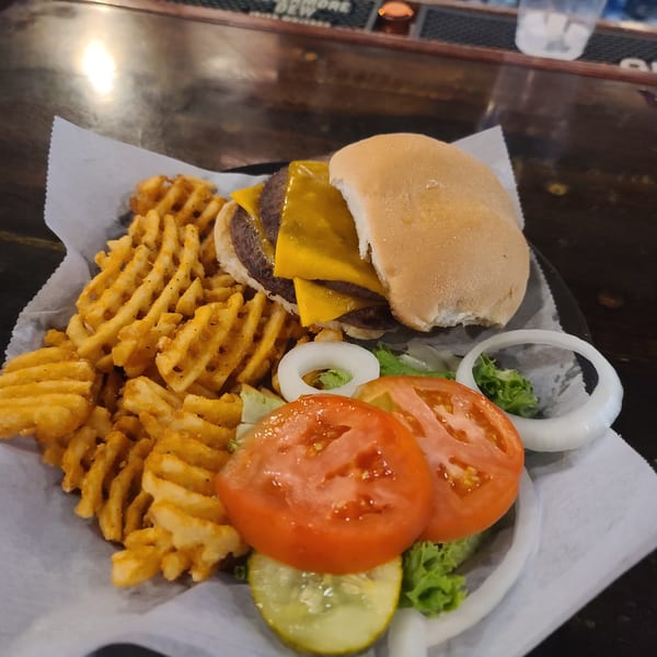 Double cheeseburger with waffle fries, lettuce, tomato, onion, and pickles on a dark bar.