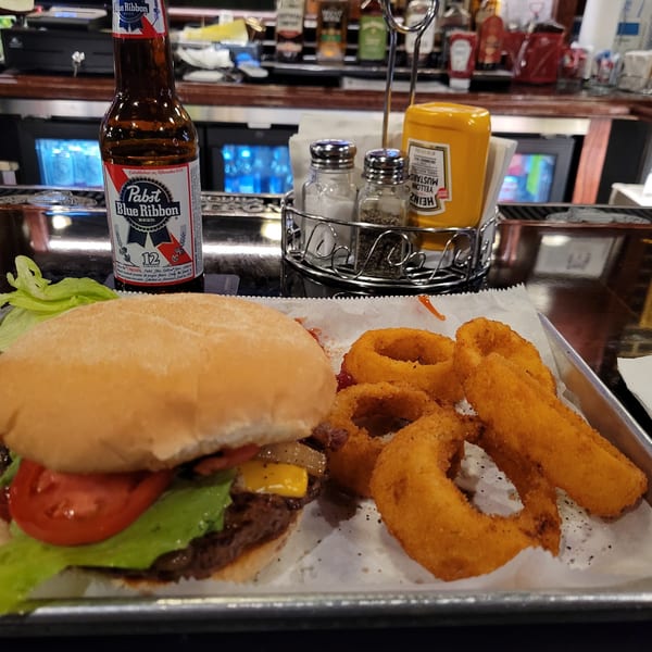 A burger with lettuce, tomato, and cheese, onion rings, and a Pabst Blue Ribbon beer on a bar.