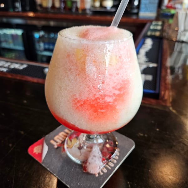 A refreshing frozen cocktail with pink and white layers, served in a stemmed glass with a straw on a bar.