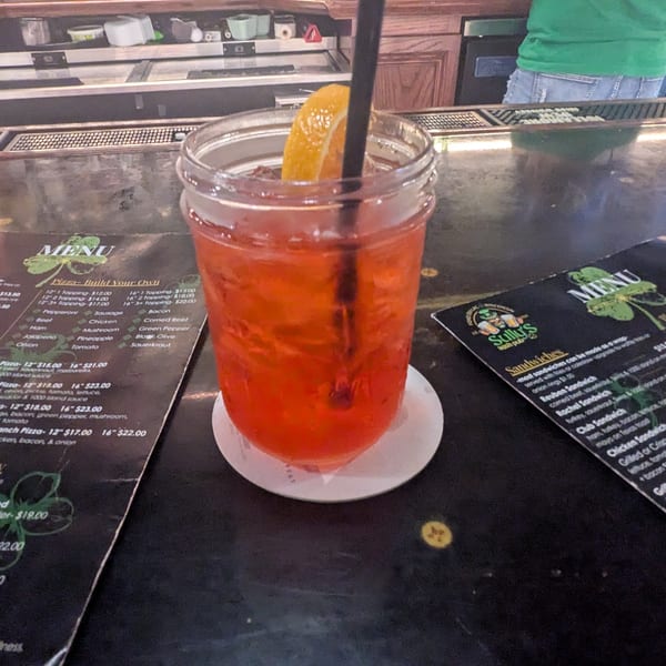 A red cocktail with an orange slice and straw sits on a bar next to menus from Sully's Irish Pub.