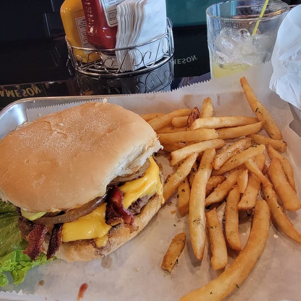 A bacon cheeseburger with lettuce, onions, and fries on a tray with a drink and condiments.