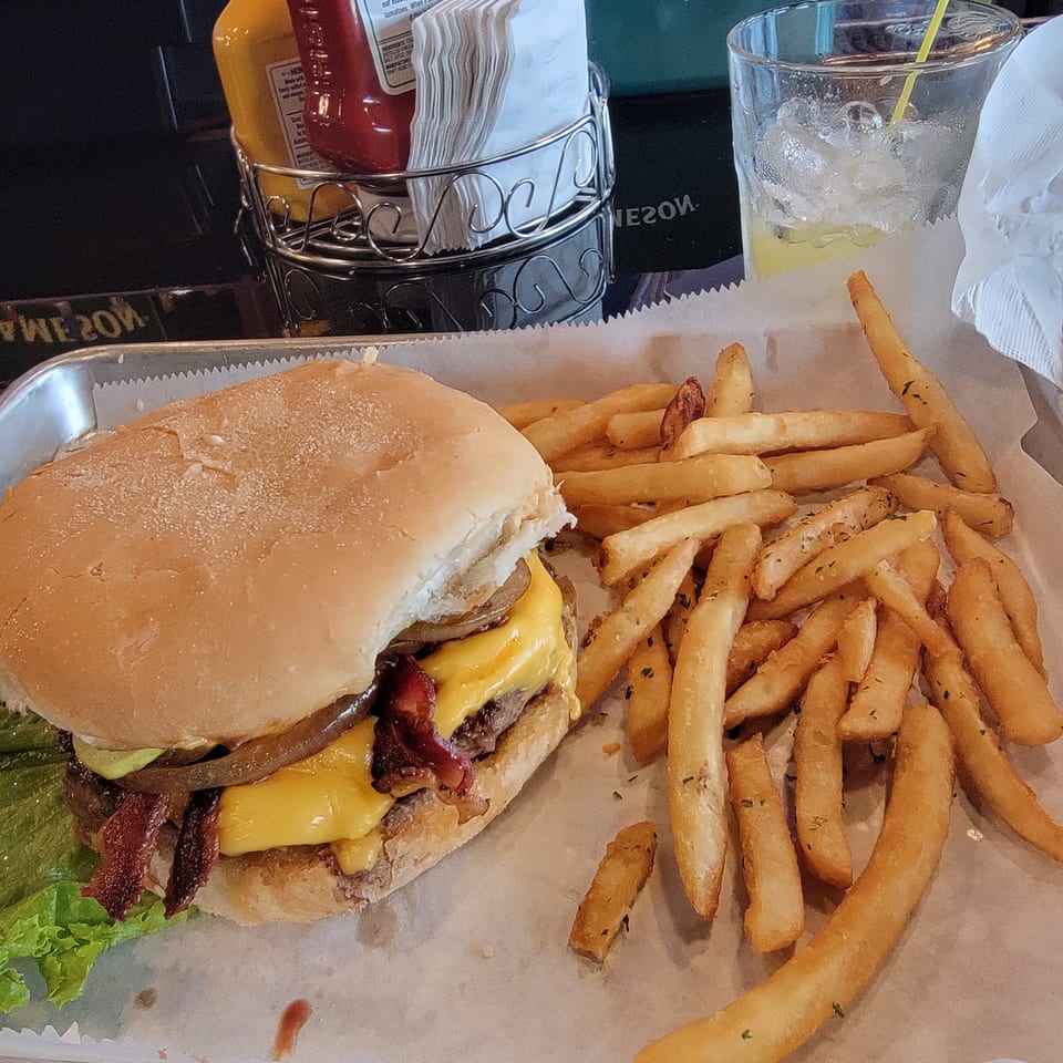A bacon cheeseburger with lettuce, onions, and fries on a tray with a drink and condiments.