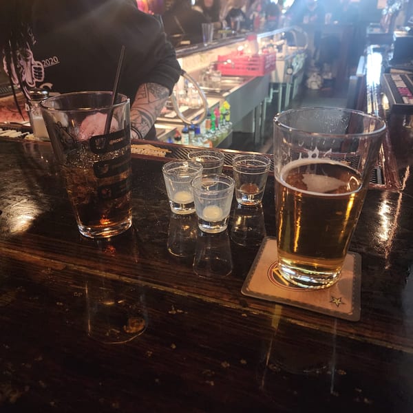 A dark bar scene with a person, drinks, and shot glasses on a wooden counter.